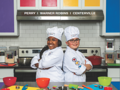 a magazine cover reads At Home in Houston county 2024. Two kids in chefs uniforms standing back to back in front of colorful kitchen background